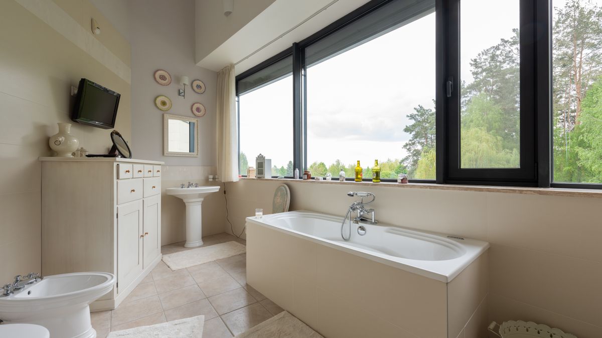 Toilet and bidet near cupboard in stylish bathroom with sink and white tub near big window overlooking green plants in tropical resort