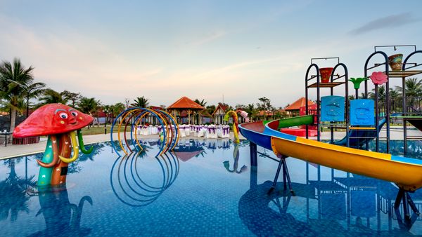 Colorful waterpark with slides, mushroom-shaped feature, and palm trees reflecting in pool.