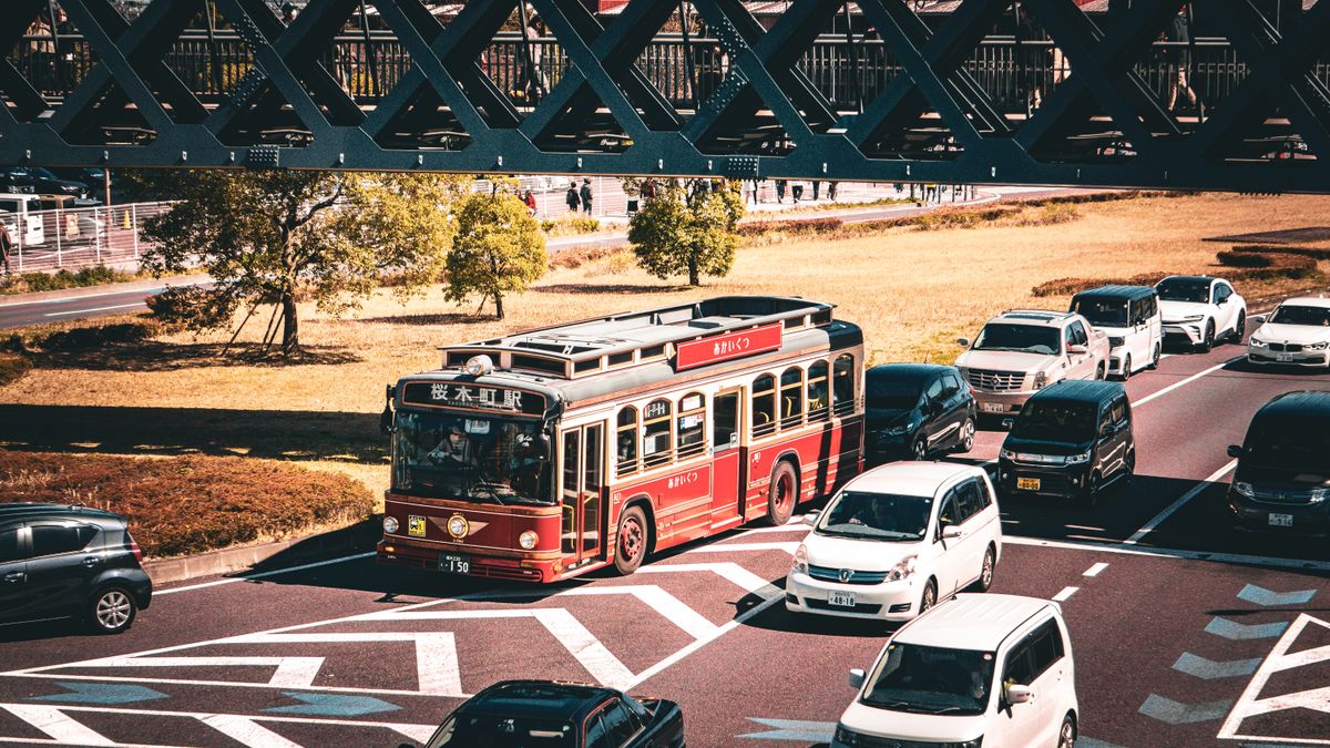 A vintage red bus travels on a busy street under a bridge, surrounded by cars.