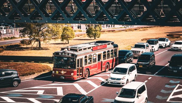 A vintage red bus travels on a busy street under a bridge, surrounded by cars.