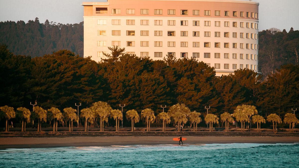 Surfer carrying board on Miyazaki Beach with hotel and sunrise backdrop in Japan.