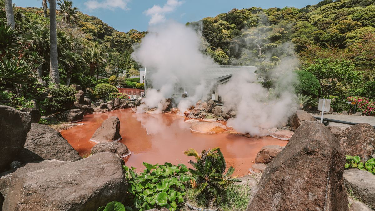 Explore a vibrant hot spring surrounded by lush greenery with rising sulfur smoke under a clear blue sky.
