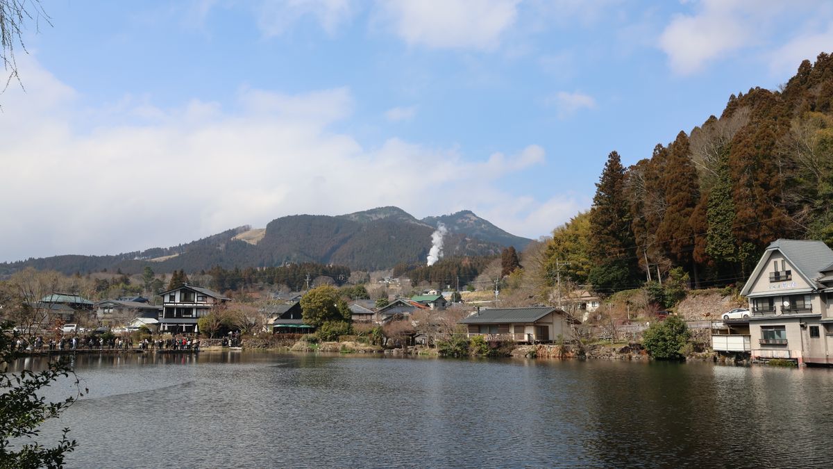 arnaud audoin / Pexels Picturesque view of Yufuin with mountains and lakeside houses under a clear sky.