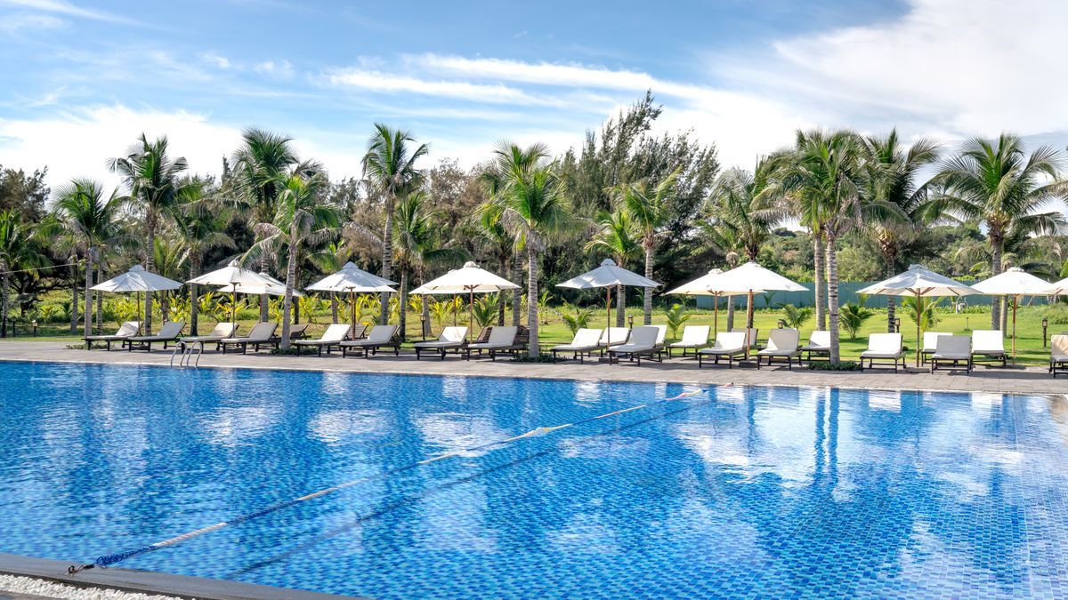 Quang Nguyen Vinh / Pexels Relaxing poolside scene with umbrellas, palm trees, and loungers under a clear blue sky.